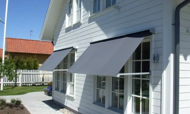 Grå markiser monterade på ett vitt trähus. Solen skiner och skapar skuggor på fasaden.