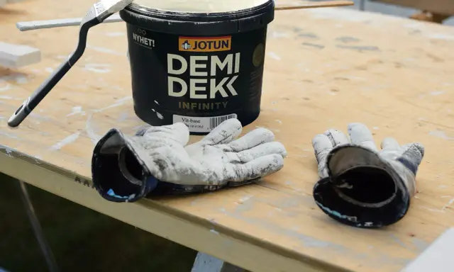 Förberedelser för målning utomhus: en burk Jotun Demi Dekk, pensel och slitna handskar ligger på ett träbord.