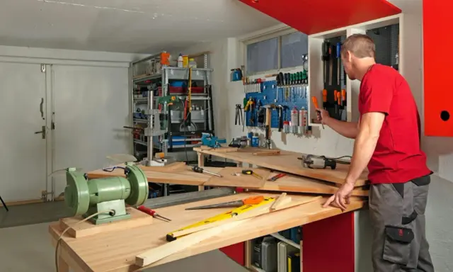 Man i röd tröja i en inredd verkstad. Verktyg på väggen, bänkar med maskiner och material.