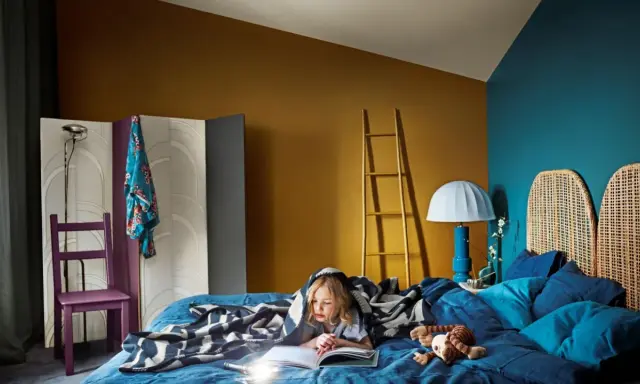 Ett barn läser under ett mönstrat täcke i ett mysigt blått sovrum. En golvlampa och en stege i trä bidrar till rummets unika atmosfär.