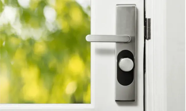 Modernt lås med nyckel på en vit dörr. Utanför syns gröna träd i oskärpa.