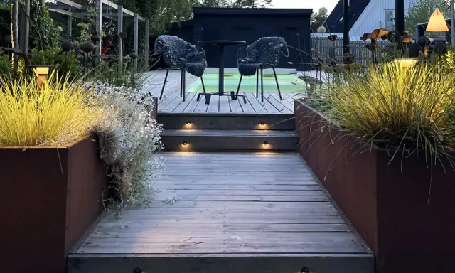 Välkomnande altan med decklights som lyser upp trappstegen. Två mysiga fåtöljer väntar vid ett runt bord.