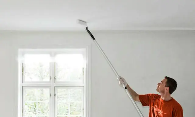 Man målar taket med en lång roller. Fönstret bakom släpper in ljus.