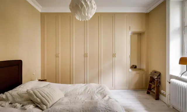 Sovrum med en stor, ljusbeige garderob som sträcker sig längs väggen. En vit taklampa med geometriskt mönster hänger ovanför.