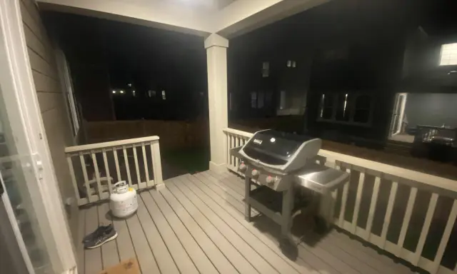 En gasolgrill under taket på altanen, redo för grillkvällar. Bredvid står en gasoltub och ett par skor.