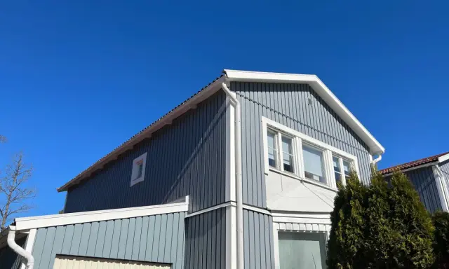 Blå himmel över ett gråblått trähus. Snart dags att måla fasad för att skydda det mot väder och vind.