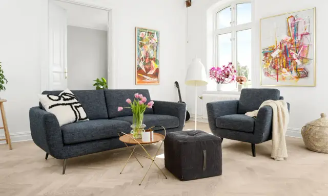 En modern soffgrupp 3+2 i mörkblått tyg, med en fåtölj, soffbord, pall och konst på väggarna.