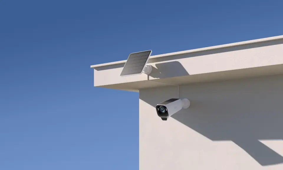 En eufycam 2 pro test visar en vit övervakningskamera med solpanel monterad på en vit vägg under en blå himmel.