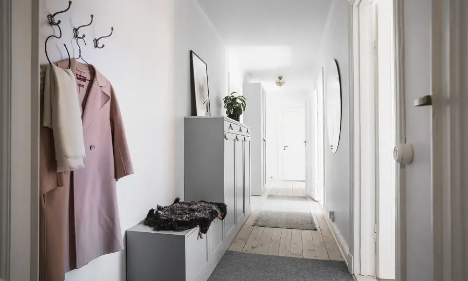 En smal hall med en rosa kappa och en vit halsduk på krokar. En grå förvaringsmöbel med en fårskinnspläd.