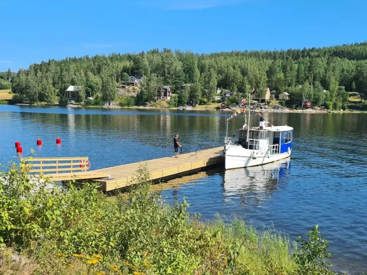 Nybyggd brygga med en b&aring;t f&ouml;rt&ouml;jd. En man st&aring;r p&aring; bryggan och njuter av utsikten &ouml;ver sj&ouml;n och de r&ouml;da bojorna.