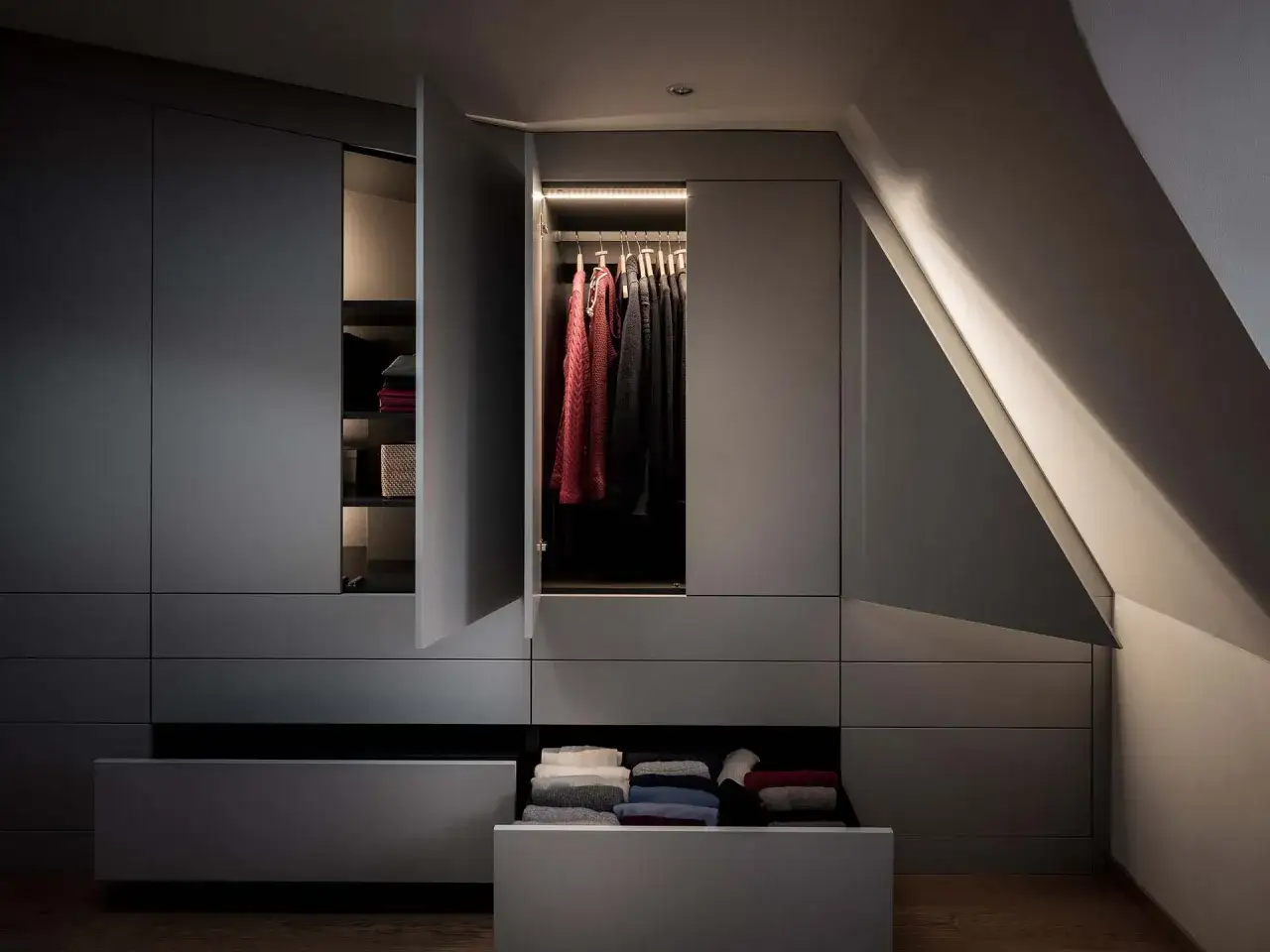 Modernt walk-in closet med snedtak. M&ouml;rka sk&aring;p med belyst kl&auml;dst&aring;ng och vikta textilier i en utdragen l&aring;da.