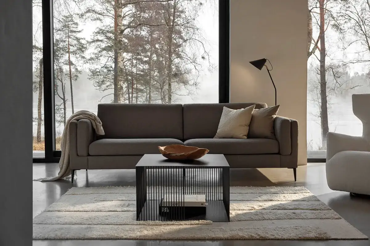 En stilren soffgrupp med en grå soffa och en svart soffbordskub, placerad framför ett stort fönster med utsikt över en skogssjö.