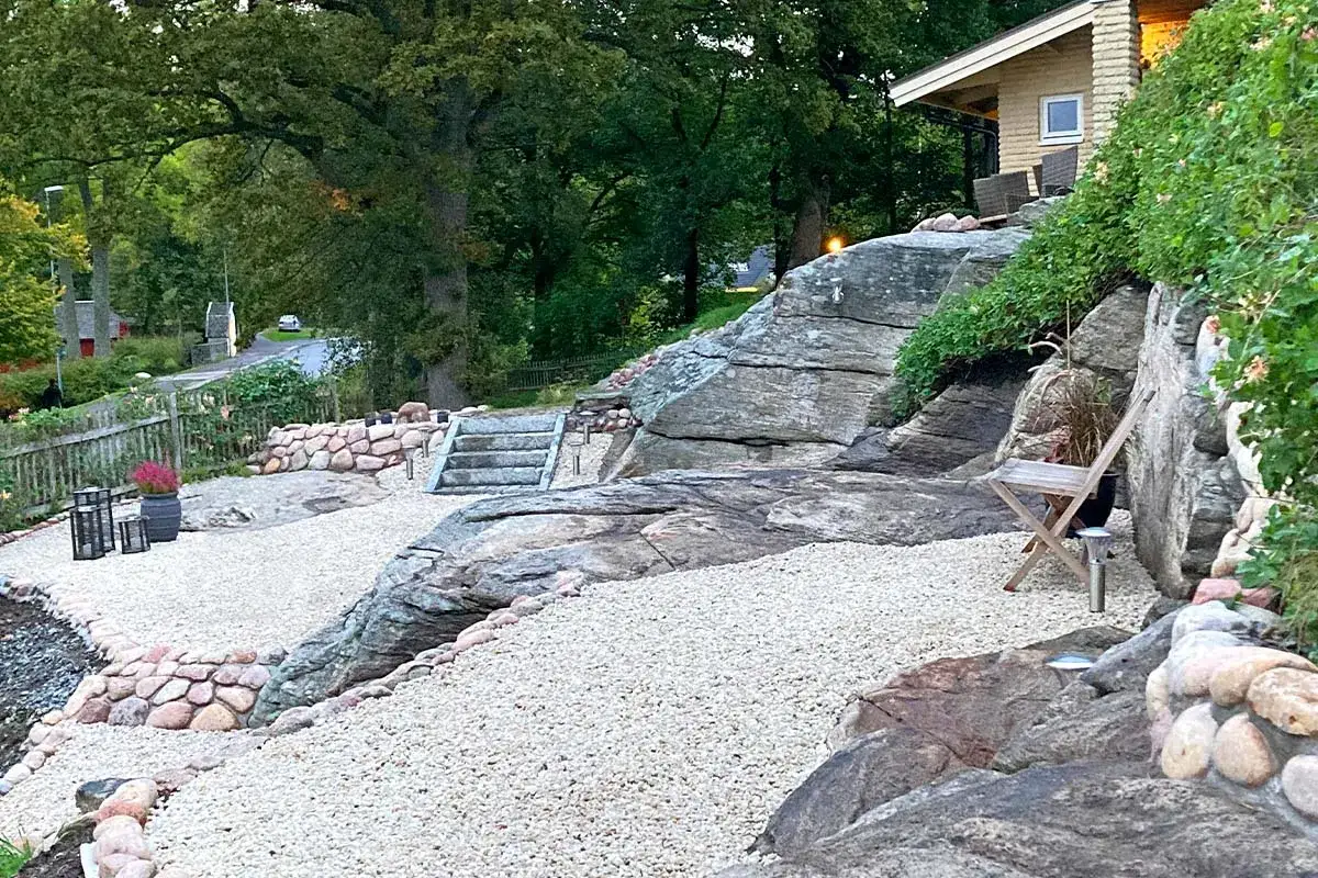 En vacker uteplats sten med grusgångar, stenmurar och en trappa som leder upp mot ett hus. En stol står redo för avkoppling.