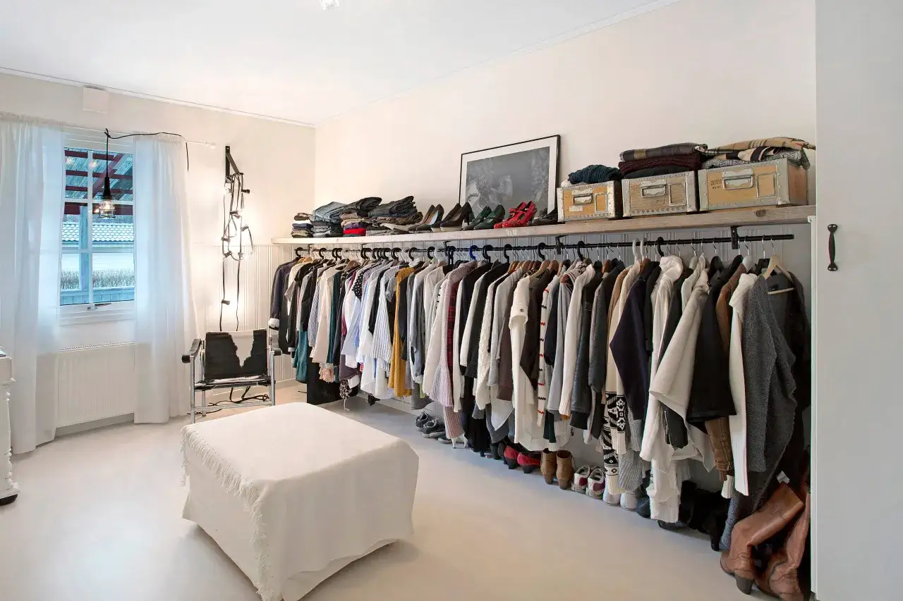 Ett stilrent, litet walk in closet med kläder på galgar, skor på golvet och förvaringslådor på hyllan. En vit ottoman står framför en fåtölj.