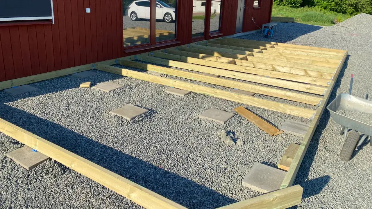 Grundarbete f&ouml;r att bygga altan p&aring; plattor. Tr&auml;reglar ligger redo att monteras p&aring; grus och stenplattor.